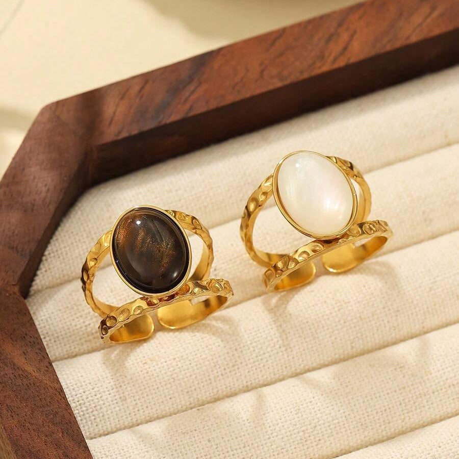 Two gold rings displayed on a cream fabric ring cushion inside a wooden box. One ring has a dark brown oval stone, and the other has a white oval stone.