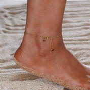 A delicate gold anklet with two dangling heart charms is shown worn on a person's ankle, set against a background of light beige sand.