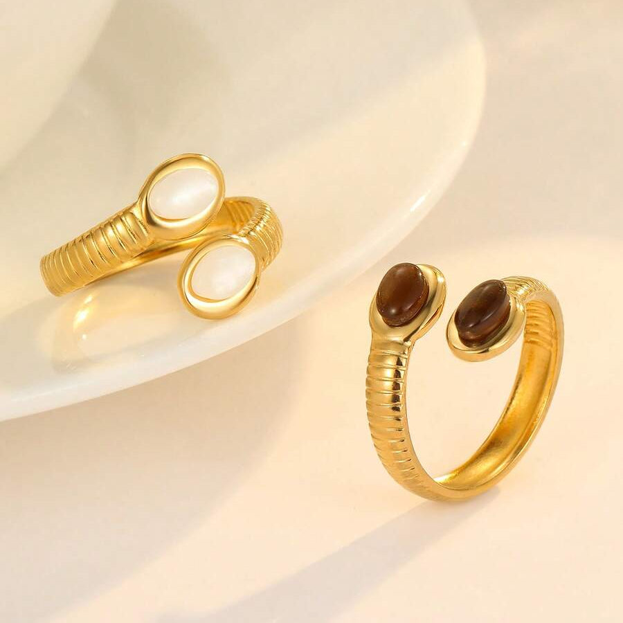 Two gold snake-themed open bangle rings displayed on a white ceramic plate and surface. One ring features a smooth white oval stone, and the other features a polished dark brown oval stone.